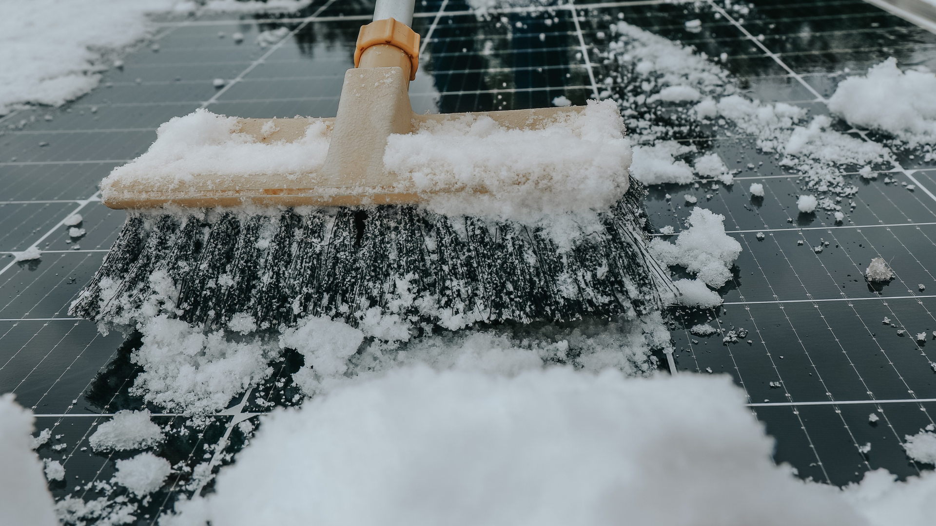 A broom pushes snow off a dark tiled surface, leaving wet patches and scattered ice.
