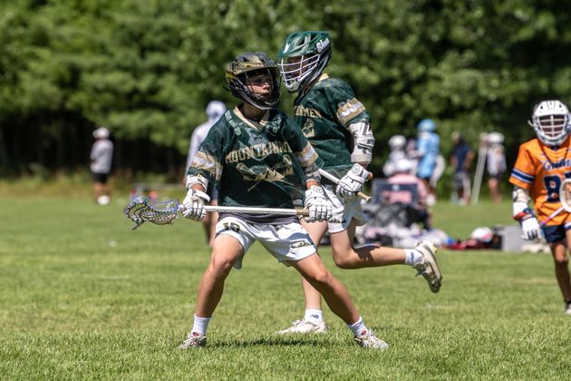 Two lacrosse players in green uniforms running on a grassy field during a game