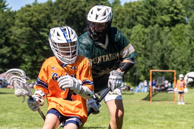 Two lacrosse players in orange and black compete on a sunny grassy field