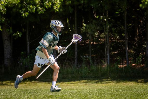 Lacrosse player in green uniform runs on grass carrying a stick, with a wooded background.