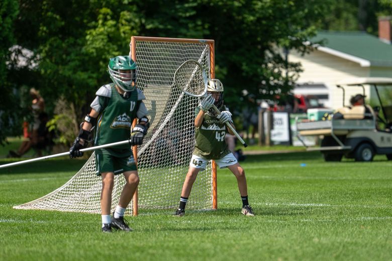 Two lacrosse players in green uniforms near a goal on a grassy field, one readying to throw