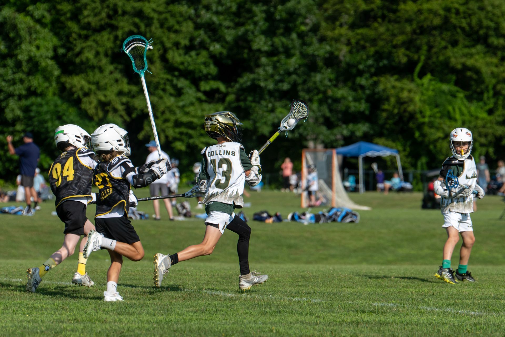 Youth lacrosse players in white and black uniforms chase the ball on a grassy field.