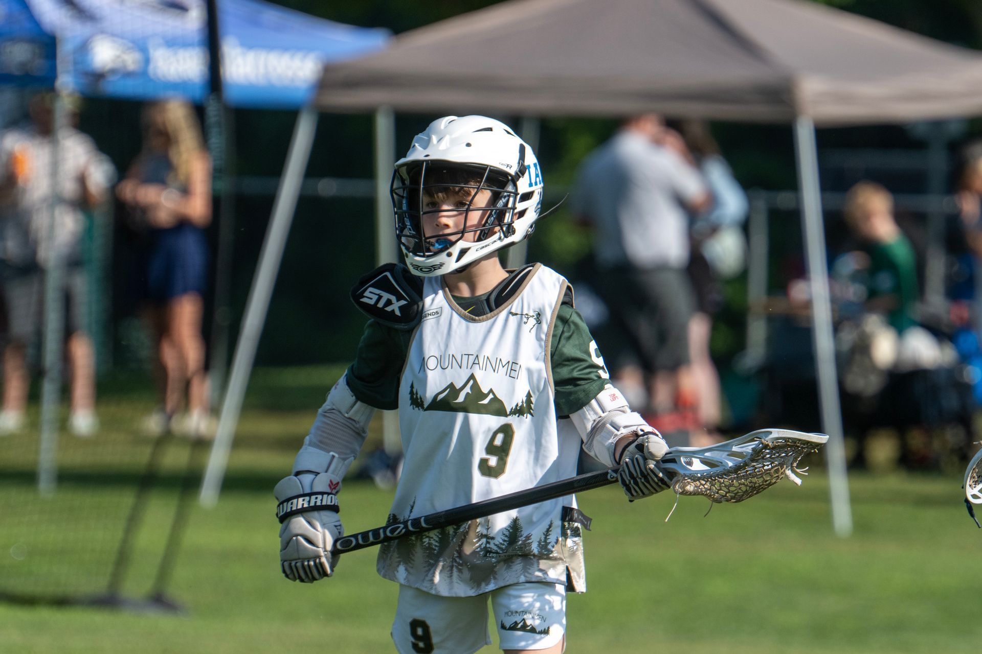 Youth lacrosse player in white uniform holding a stick on a grassy field under tents