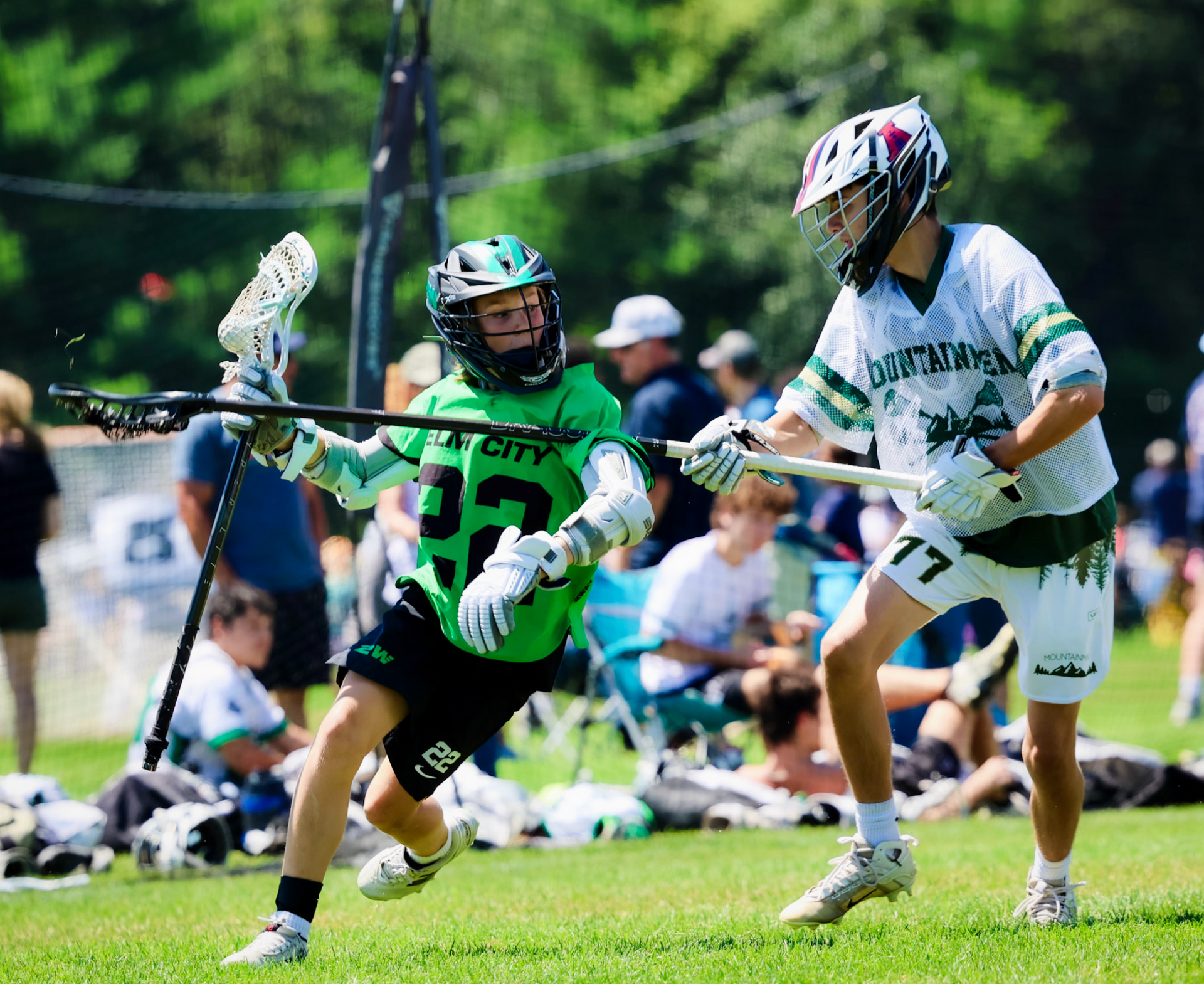 Two lacrosse players in green and white battle for the ball on a grassy field.