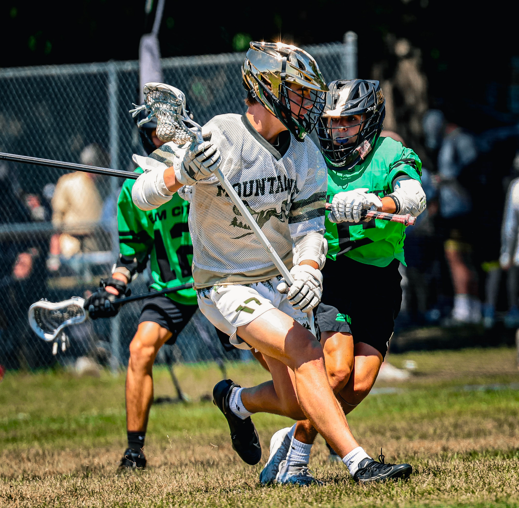 Lacrosse players in green and white uniforms sprint and jostle for the ball on a sunny field.