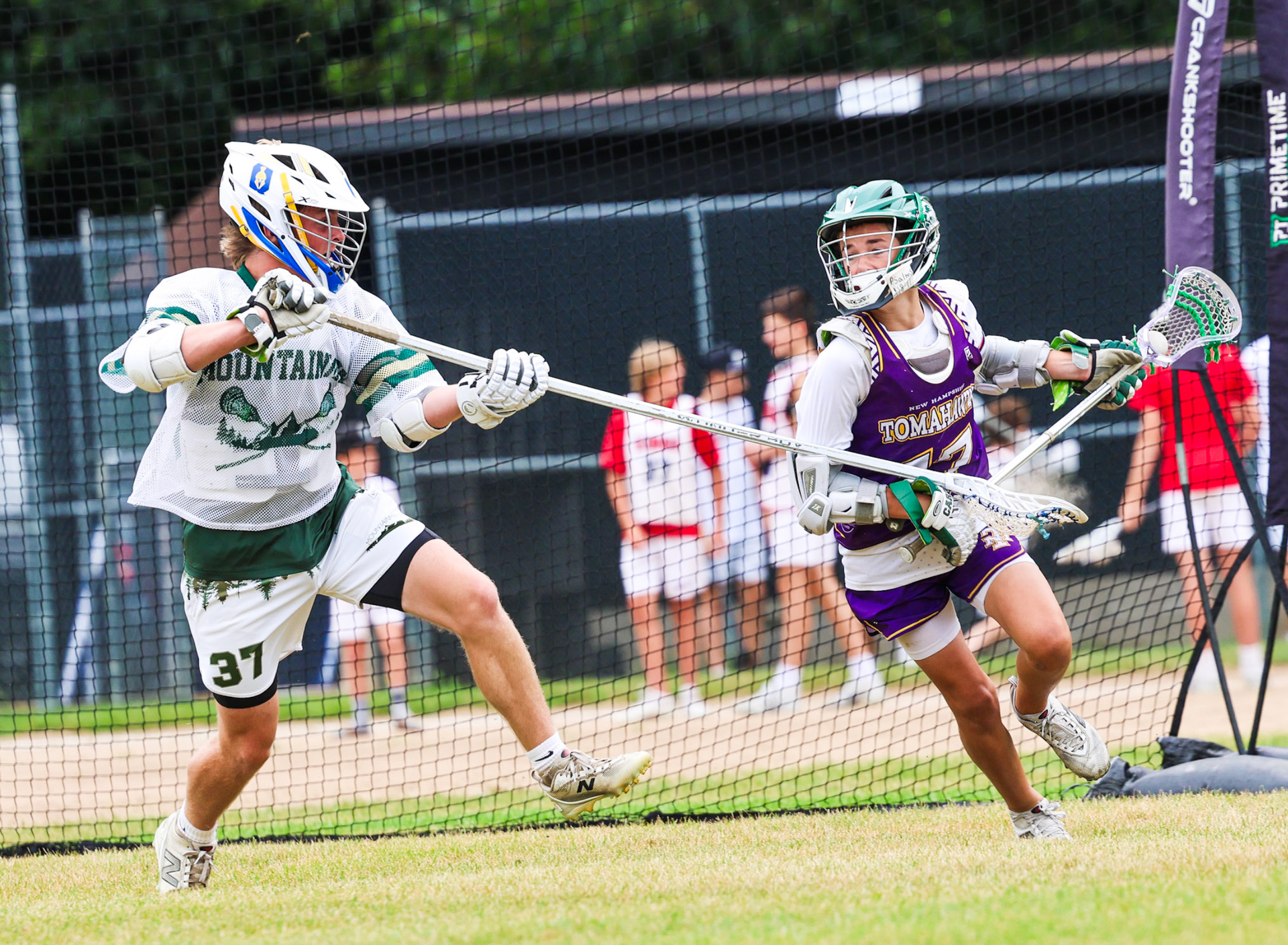 Two lacrosse players sprint and battle for the ball on a grassy field near the goal.