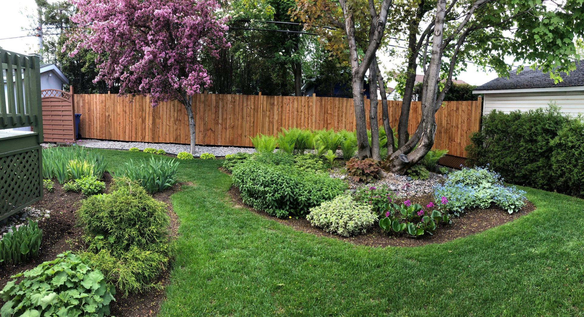 Jardin arrière luxuriant et verdoyant avec allée sinueuse, parterres de fleurs, clôture marron et arbres.