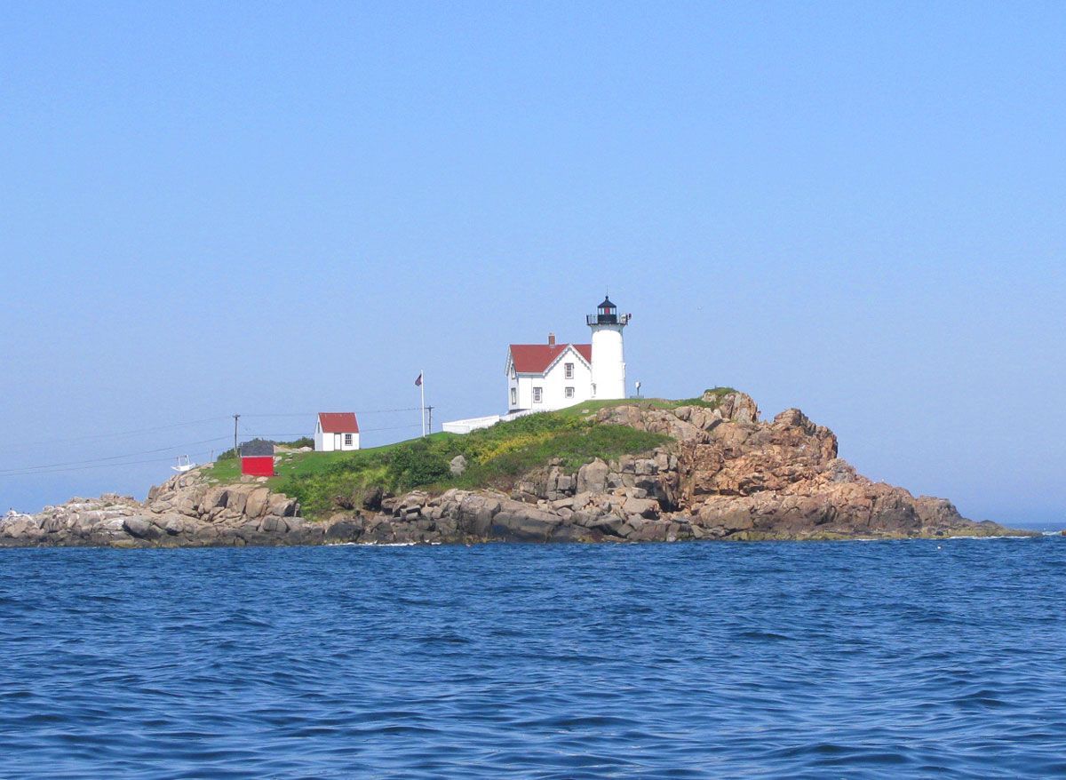 Lighthouse on a rocky island, white with a black top, red roofed buildings, blue water and sky.