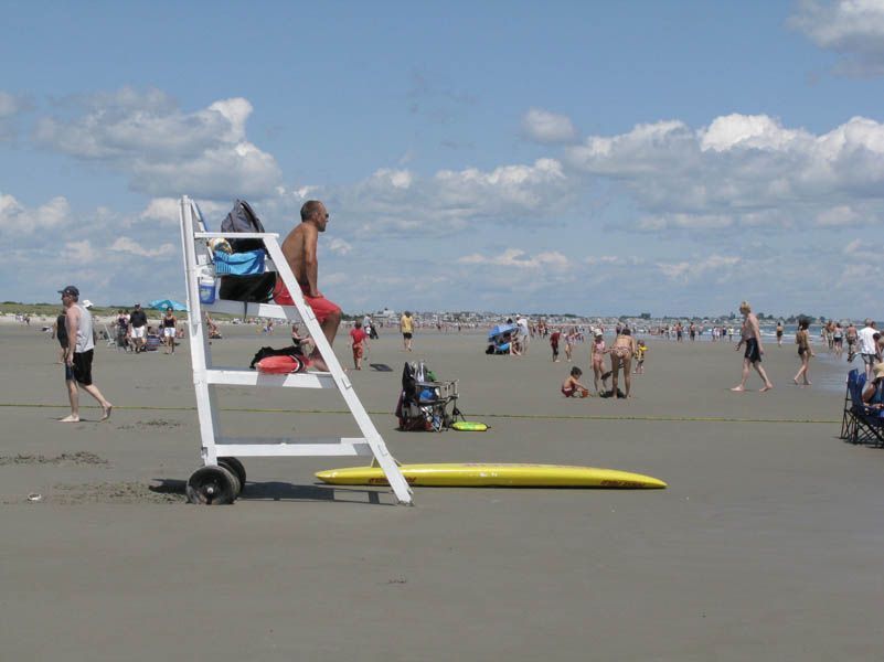 Lifeguard in red trunks on high chair, beach with many people on a sunny day.