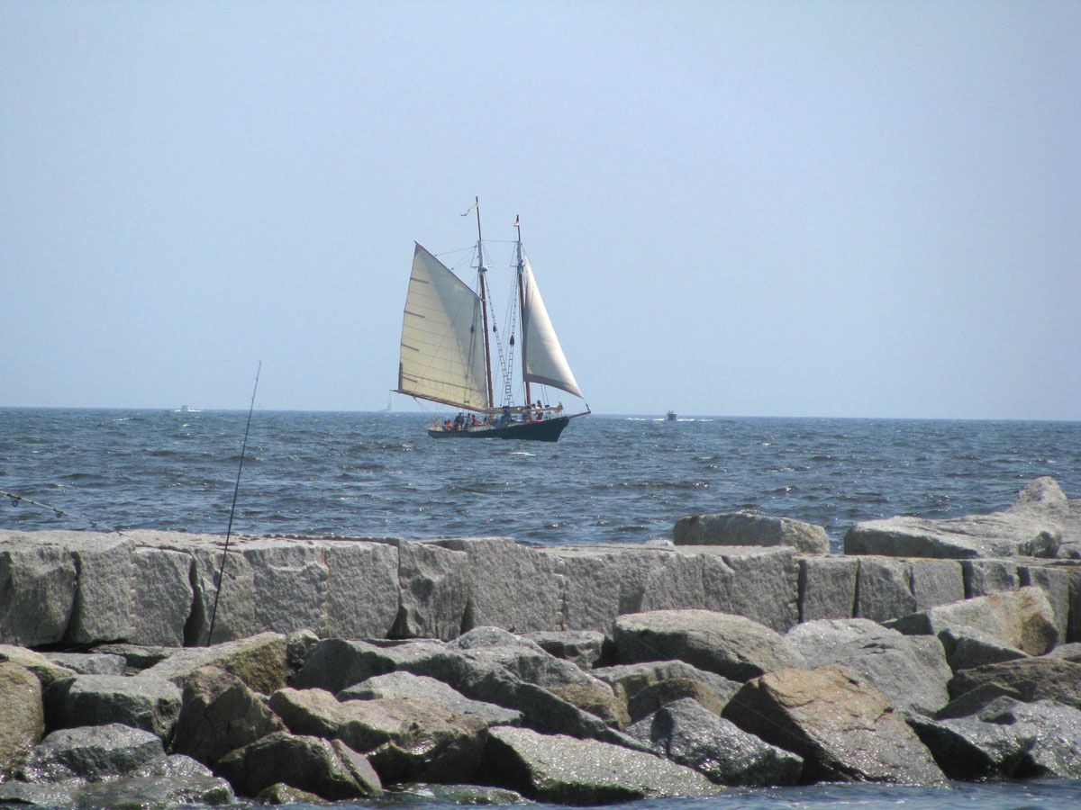 Sailboat on a blue sea, sailing past a rocky breakwater on a sunny day.