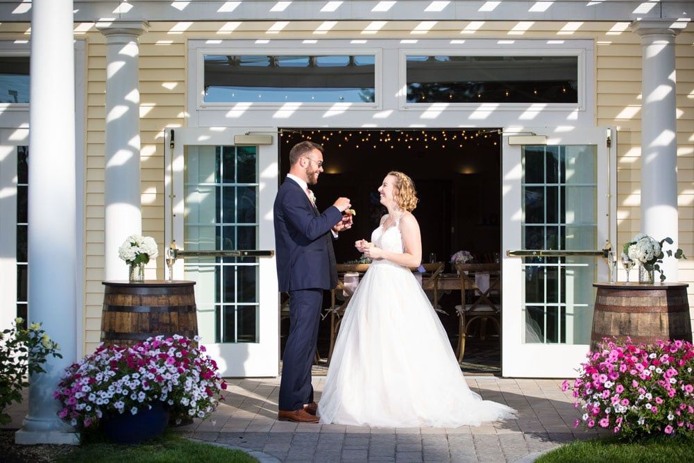Bride and groom toast with glasses, standing in front of an open building's doors, flowers surround.