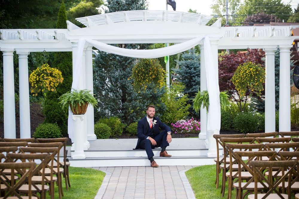 Groom in navy suit sits on white altar steps, surrounded by garden. Brown chairs line path to him.