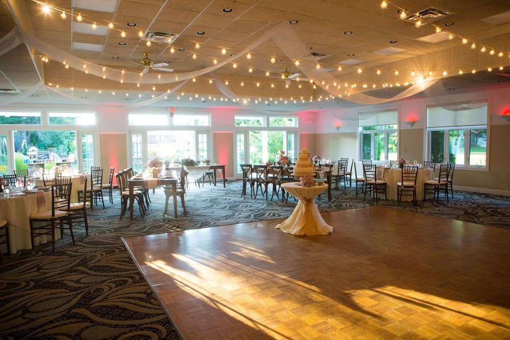 Wedding reception hall with string lights, tables, and a cake on a stand. Windows and dance floor.