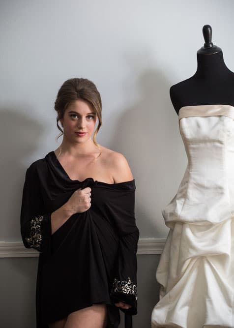 Woman in black wrap, looking at the camera, next to a wedding dress on a mannequin.