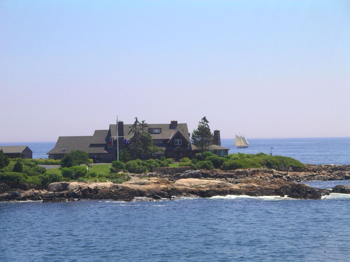 Large house on rocky island, blue ocean and sky. Sailboat in distance.