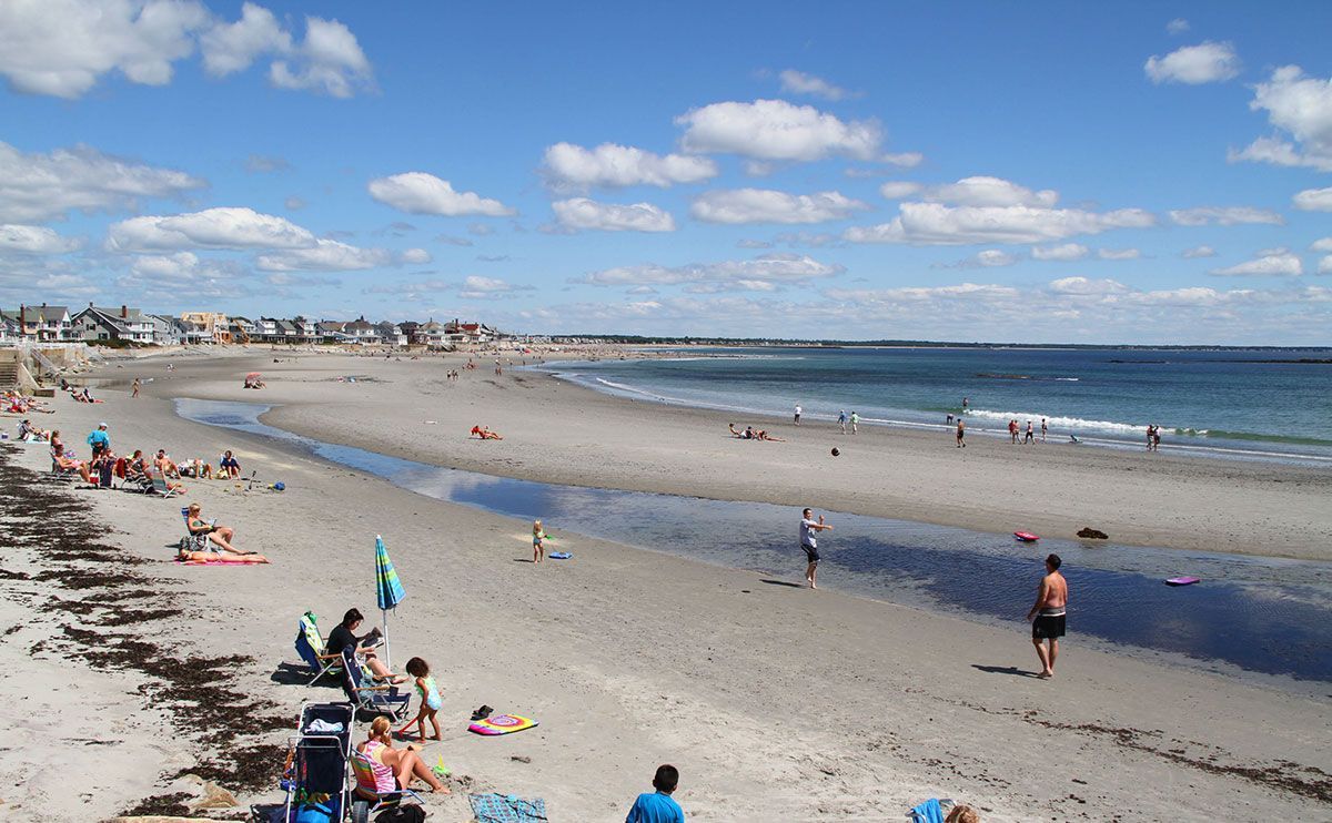 Beach scene: People on sandy beach, blue water, cloudy sky, oceanfront houses in the distance.
