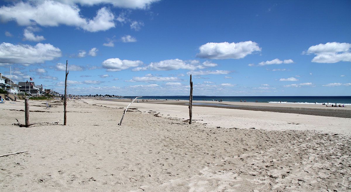 Sandy beach with volleyball net posts under a bright blue sky with scattered clouds.