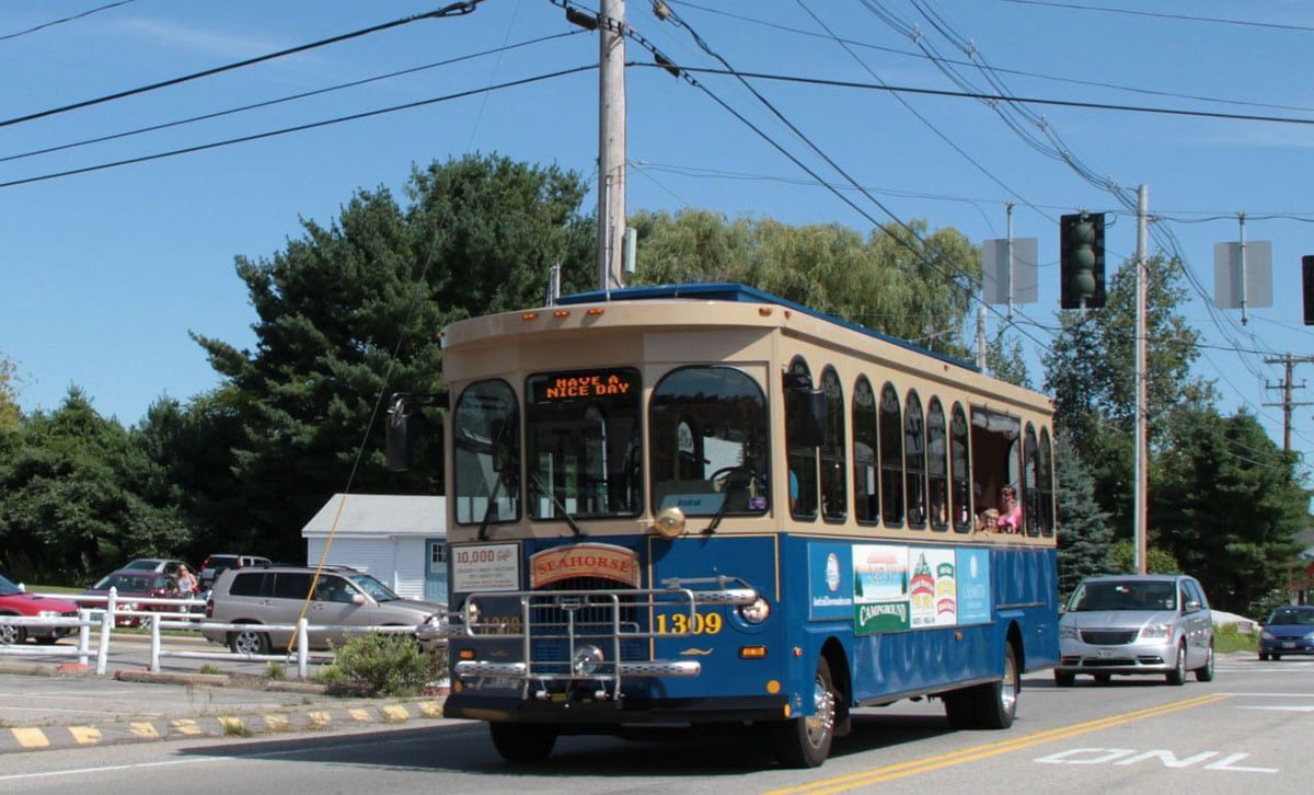 A blue trolley bus on a sunny road. It has a tan top and is near power lines and other cars.