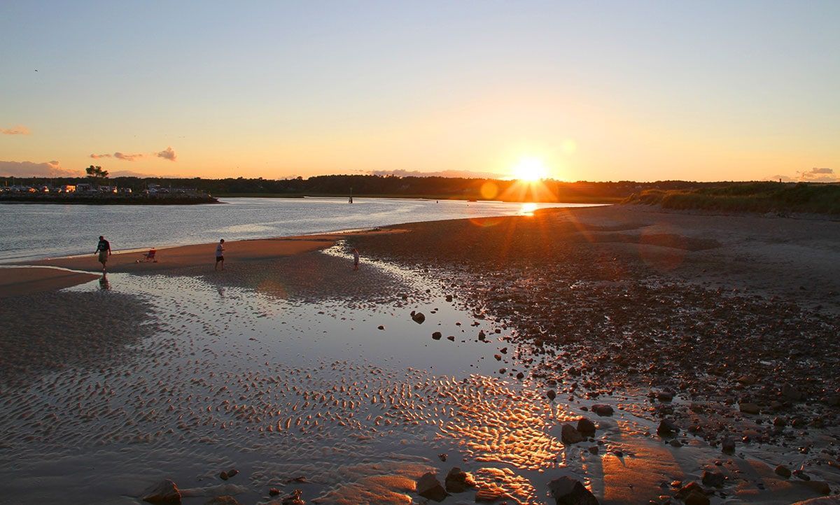Sunset over a shallow shoreline with people walking. Orange and yellow sky reflecting in water.