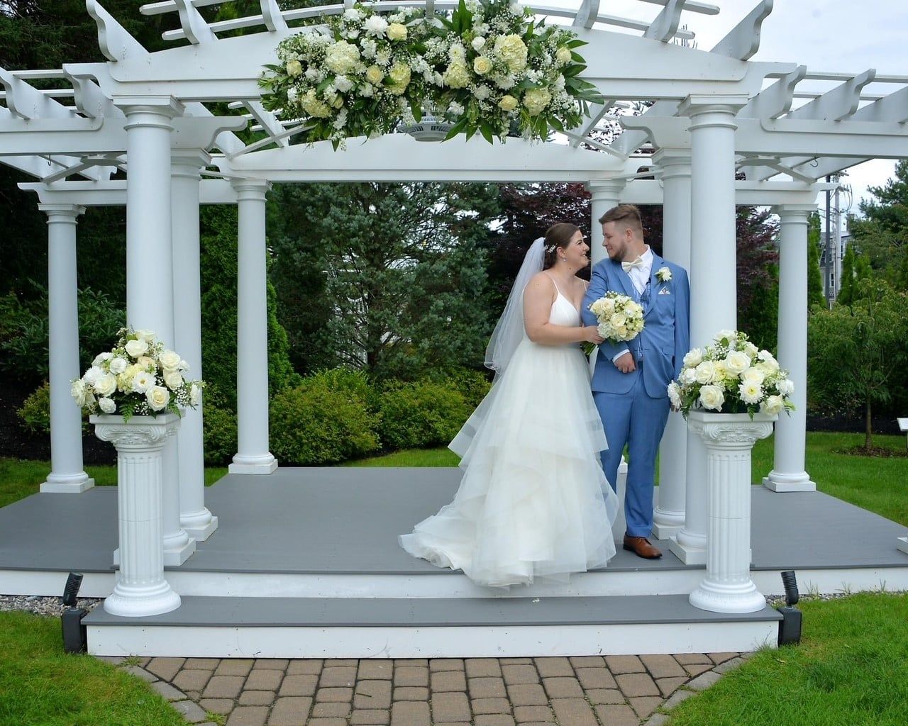 Bride and groom pose under a white floral decorated arbor, smiling. She wears a white dress; he wears a blue suit.