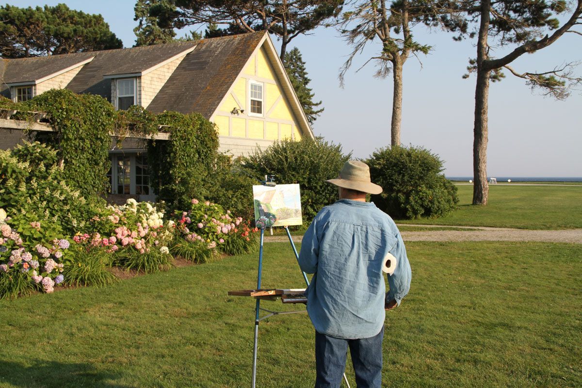 A person in a hat paints outdoors in front of a house with a green lawn, flowers, and the ocean.
