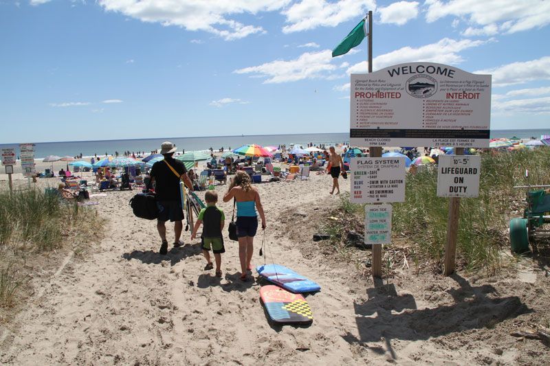 Family walking on sandy path toward crowded beach; sign reads 
