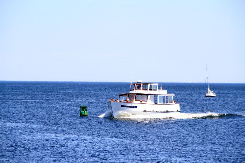 White boat on blue water with a green buoy in foreground and sailboat in the distance, under a clear blue sky.