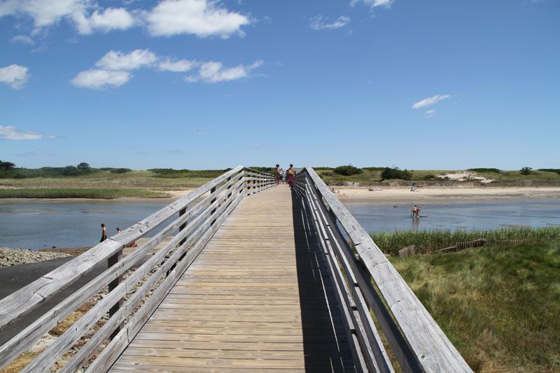 Wooden boardwalk over water to a beach; people visible on the sand and walking. Sunny day, blue sky.