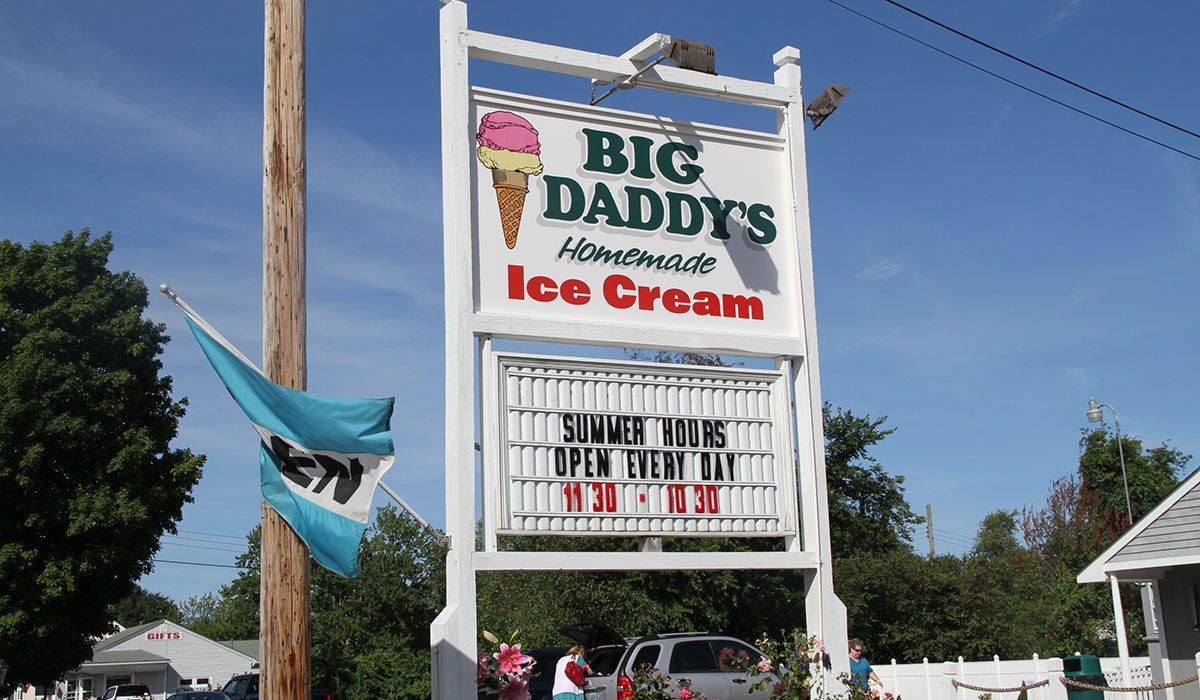 White sign for Big Daddy's Homemade Ice Cream with hours listed, blue flags, and sky.