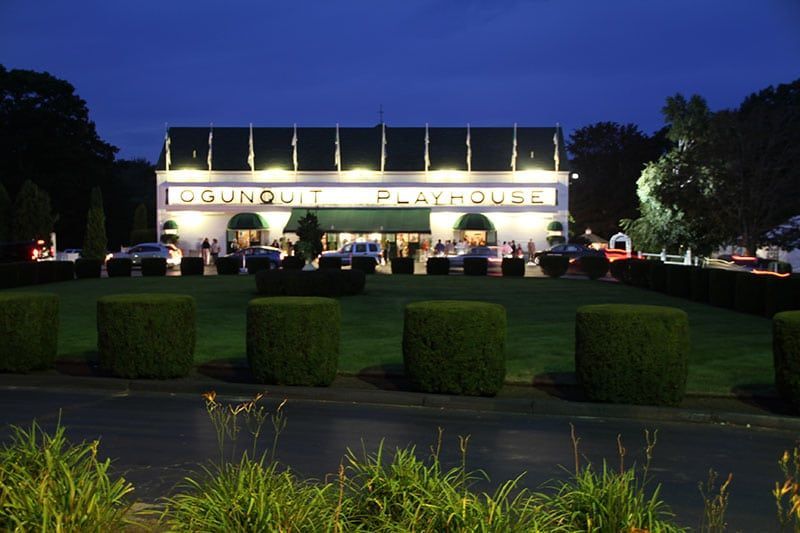 Dusk view of the Ogunquit Playhouse, a theater with lit marquee, lawn, and shrubs. Cars and trees flank the building.