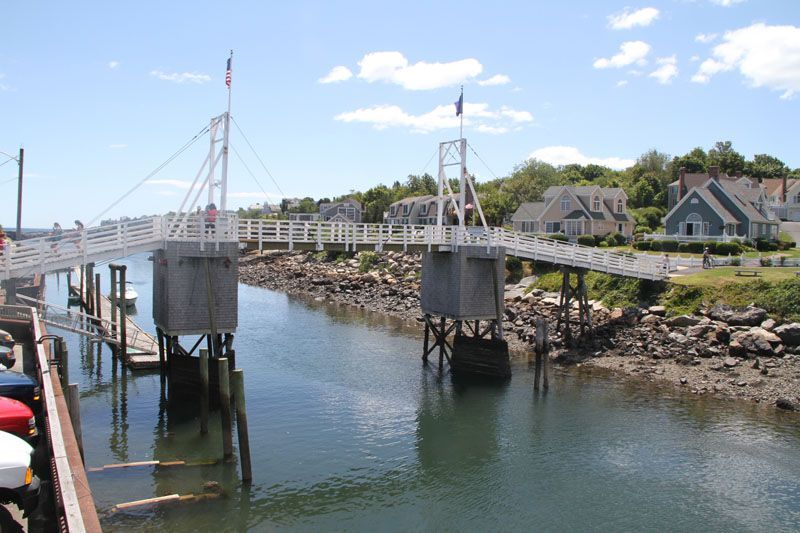 White pedestrian bridge over a harbor, with flags, wooden piers, and houses in the background.