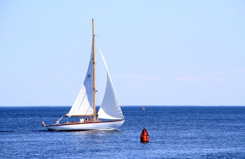 Sailboat sailing on blue water, under a clear blue sky. A red buoy is nearby.