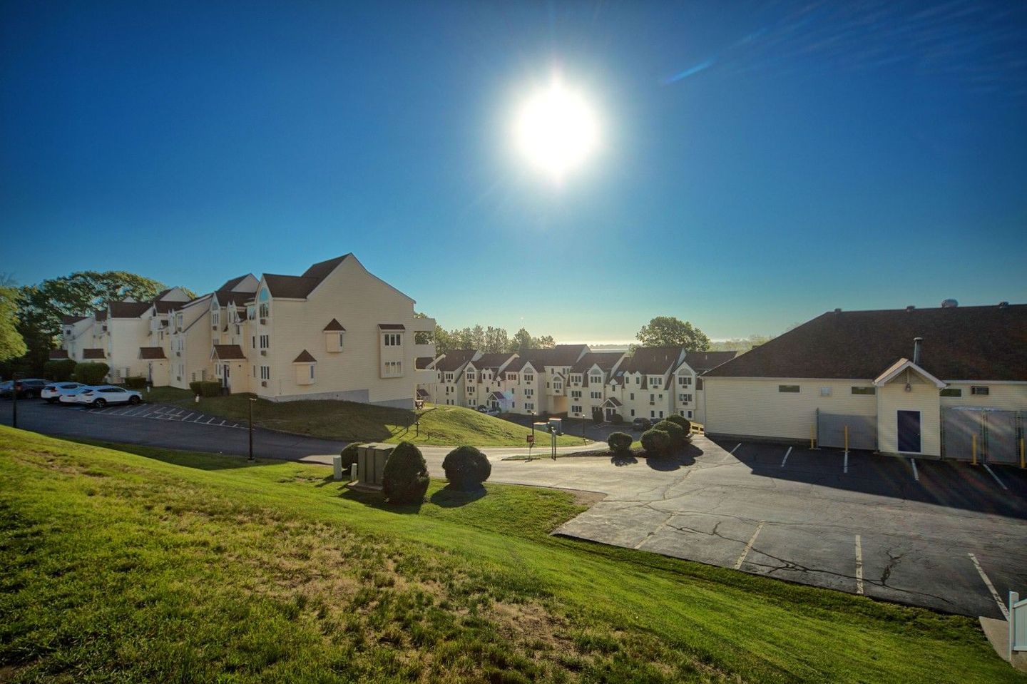 Bright sun over white buildings on a grassy hill, with a clear blue sky.