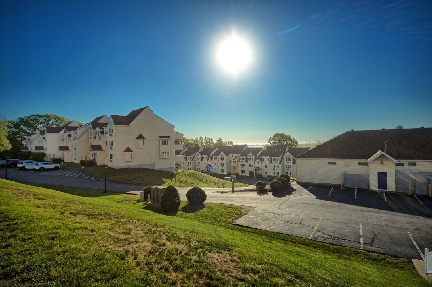 Bright sun over white buildings on a grassy hill, with a clear blue sky.