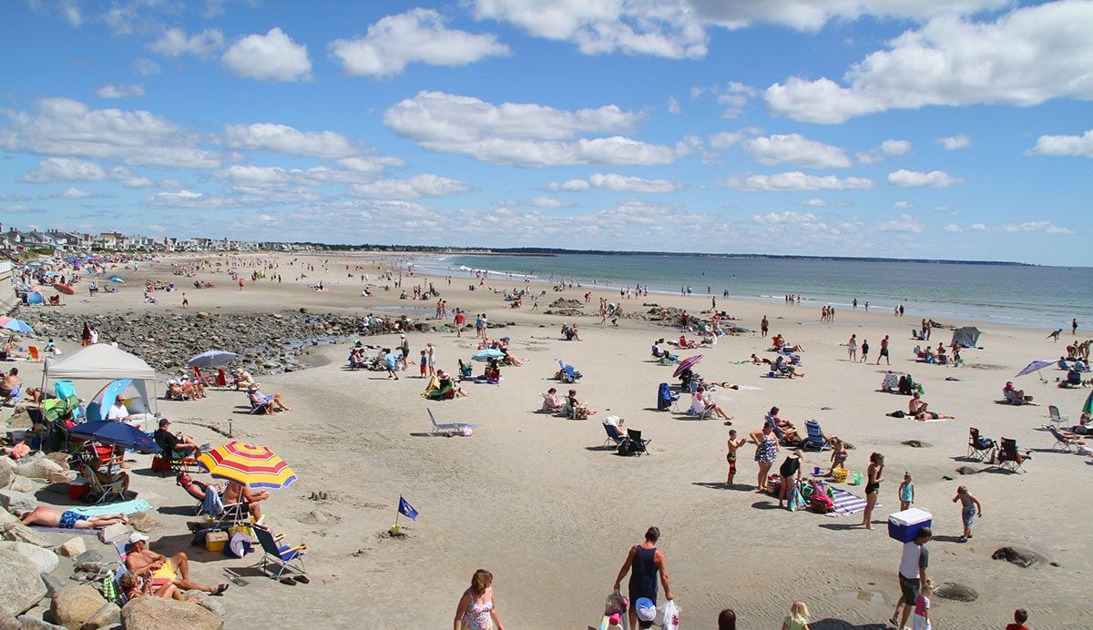Beach scene: Numerous people on a sandy shore on a sunny day with blue sky, white clouds, and ocean in background.