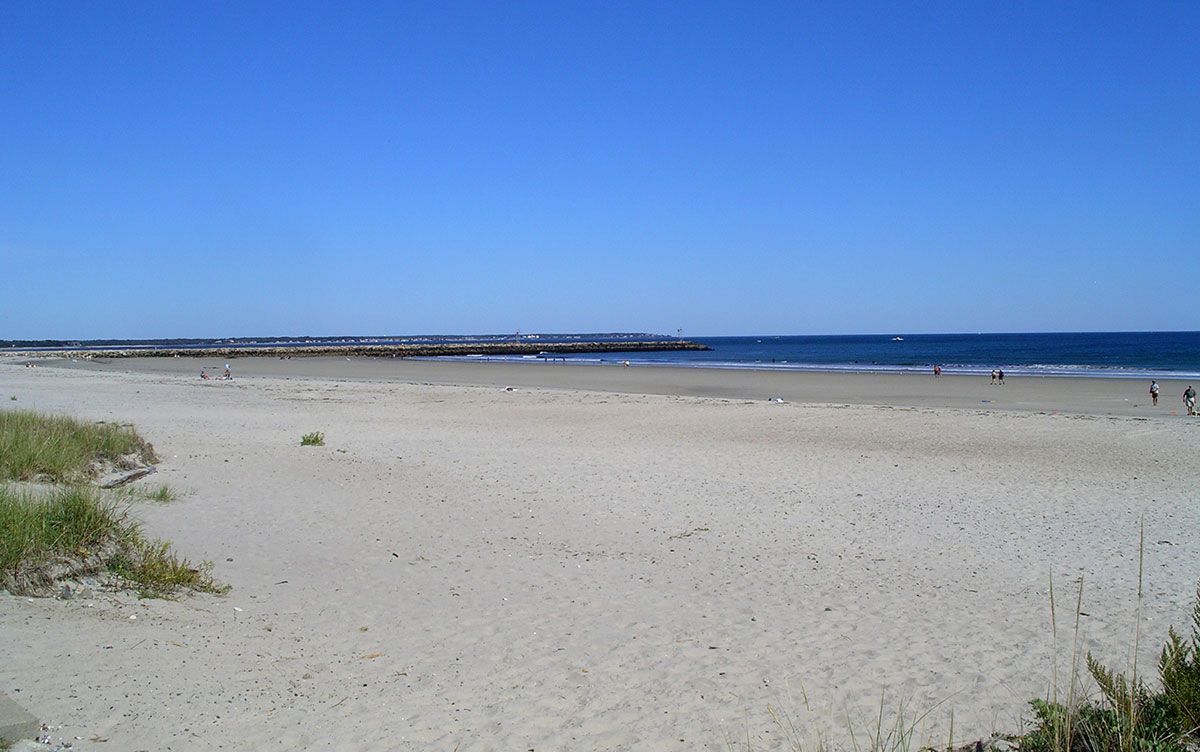 Sandy beach under a bright blue sky. Ocean waves in background with a jetty in the distance.
