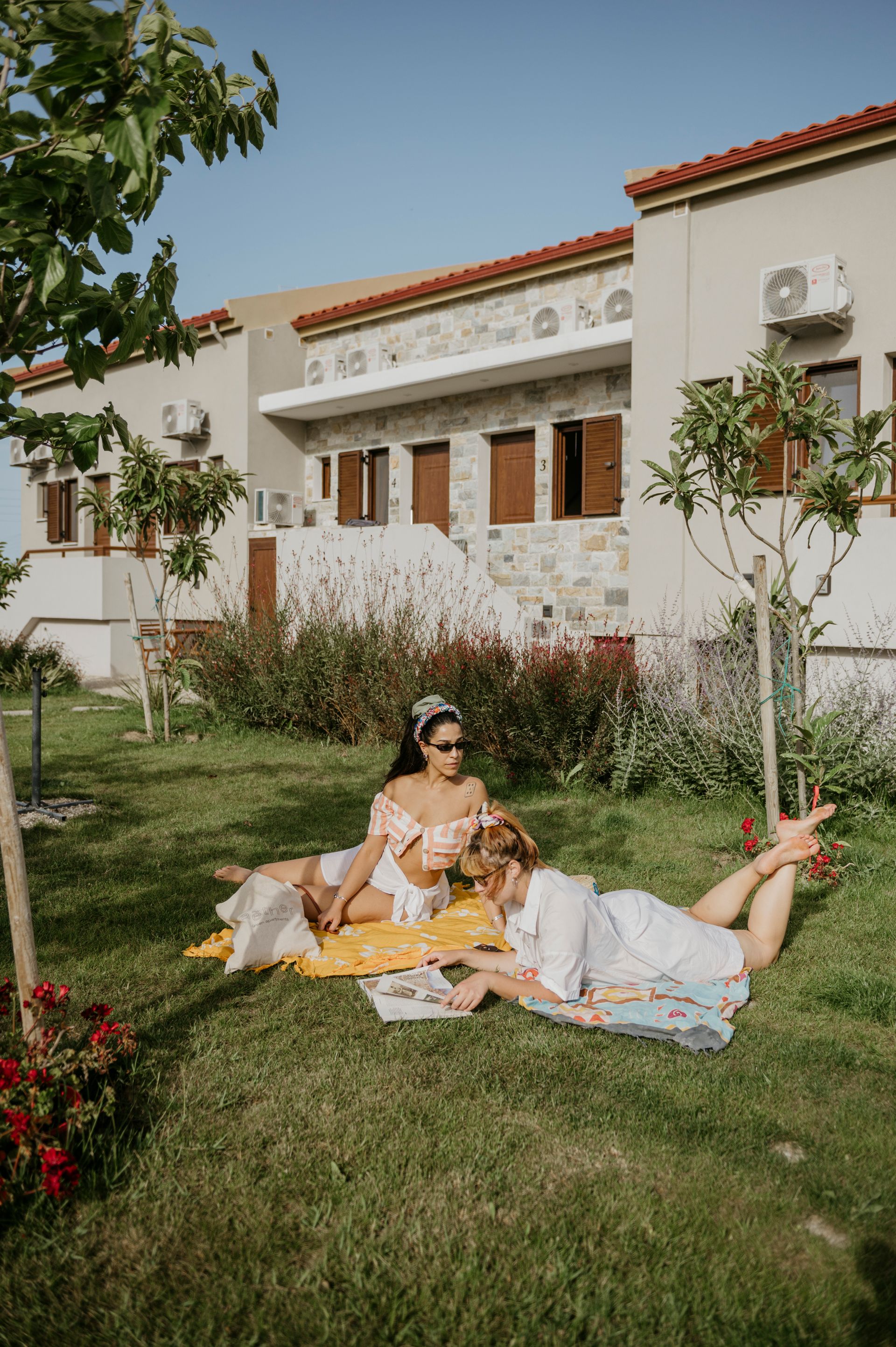 Two women are laying on a blanket in the grass in front of a building.