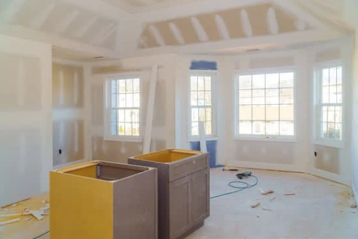Unfinished room with white walls, large windows, and kitchen cabinets in the center, under renovation