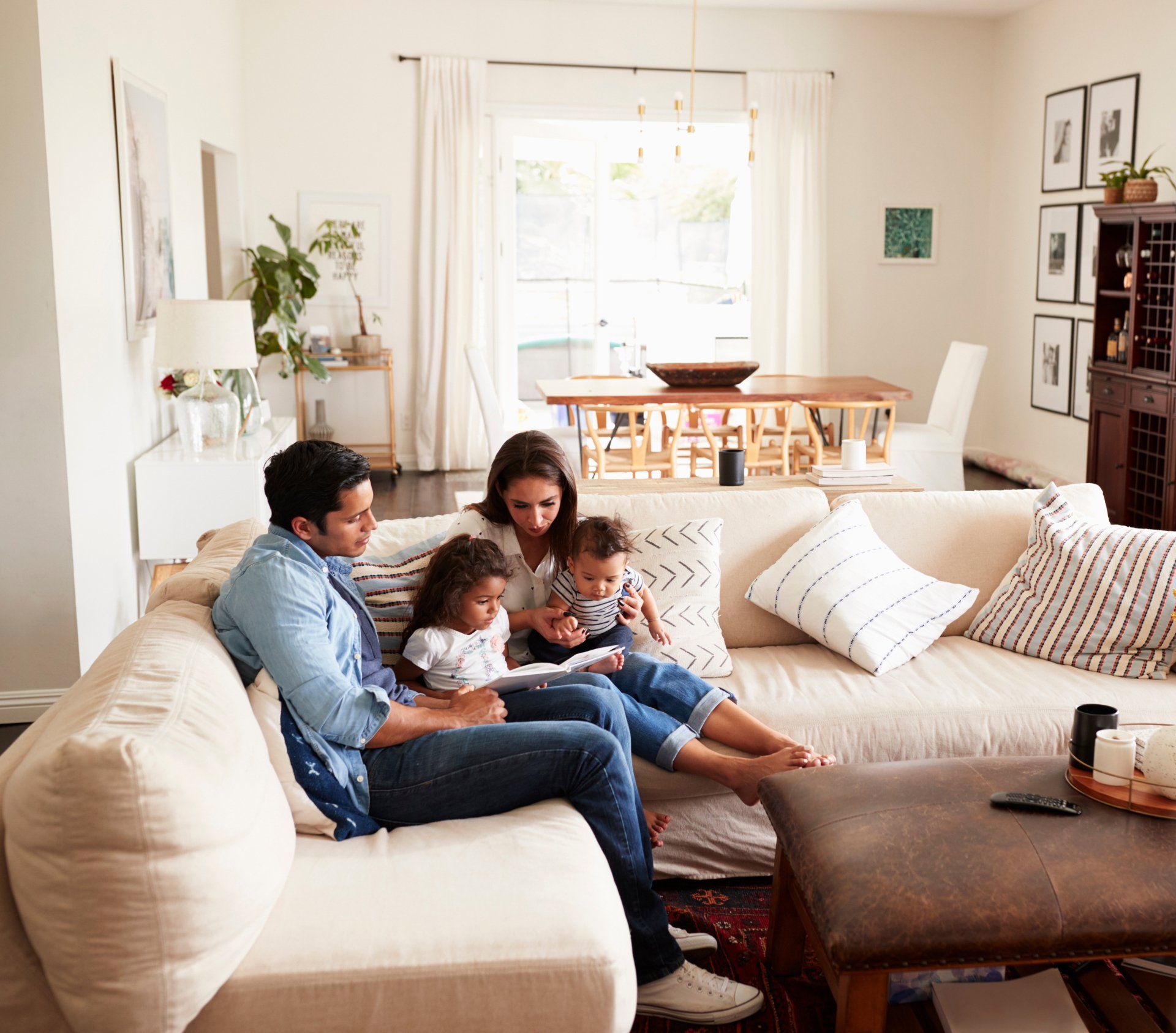 Young Hispanic family of four sitting on sofa reading a book together in the living room.