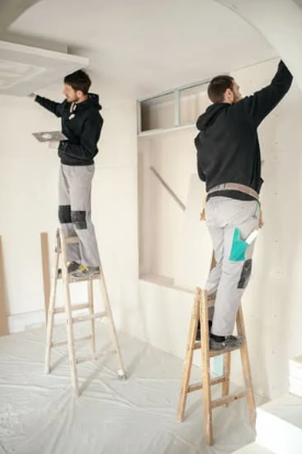 Two workers on step stools painting a white interior wall during home renovation