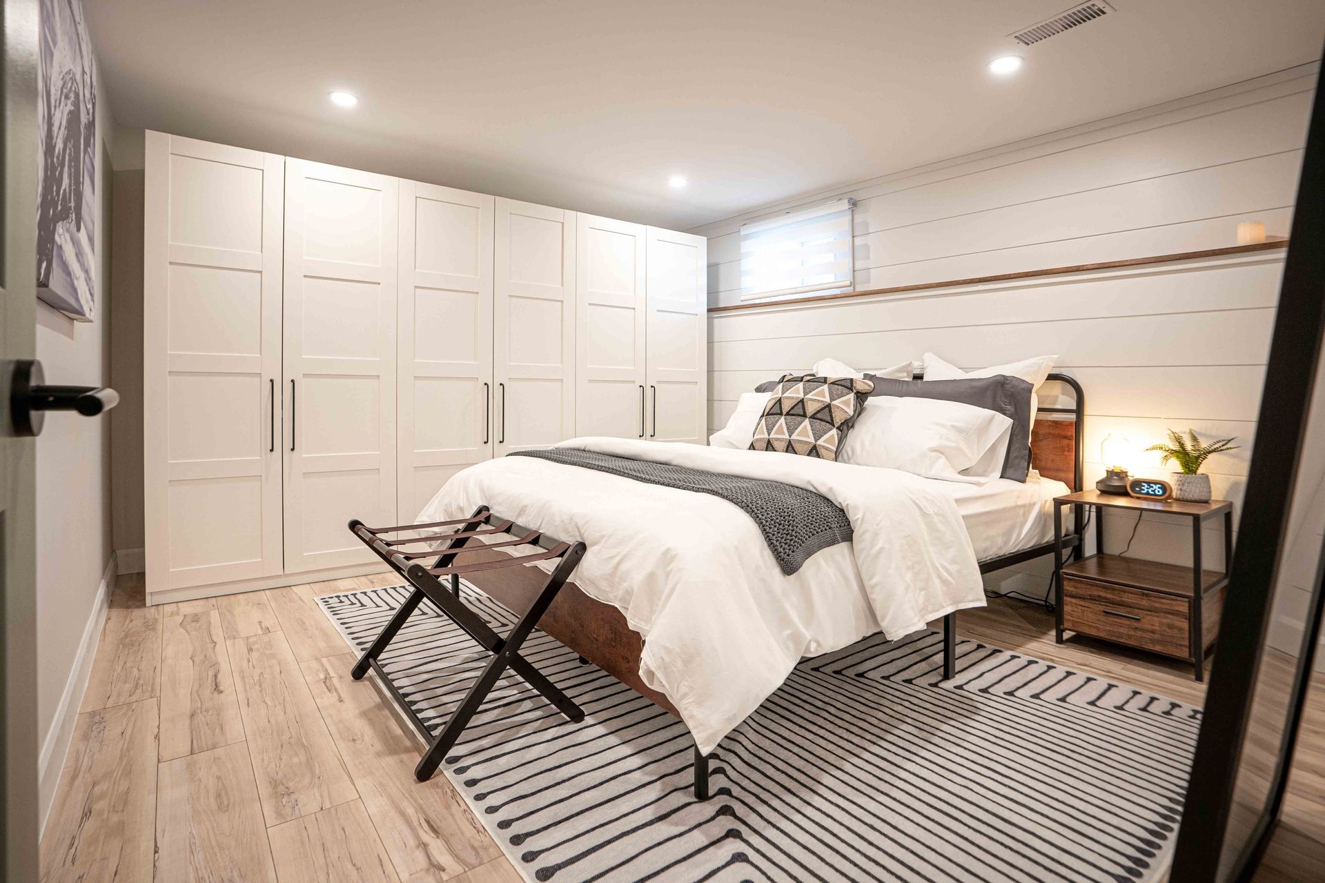 bedroom with light brown floors, a black and white rug, large white accesory closet in the back ground, white shiplap walls