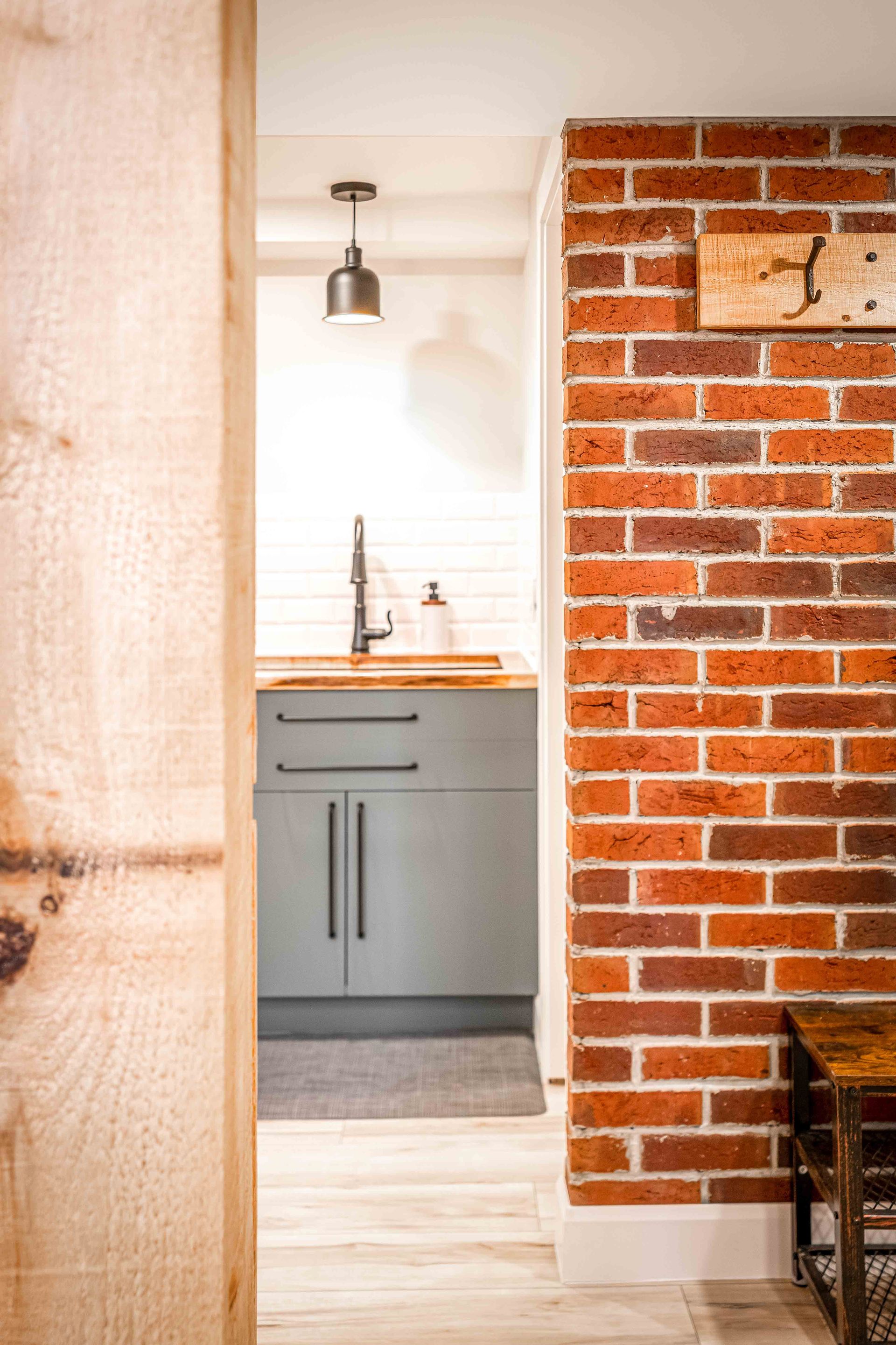 kitchen sink with light green cabinets, red brick accent wall with coat hangars and bench