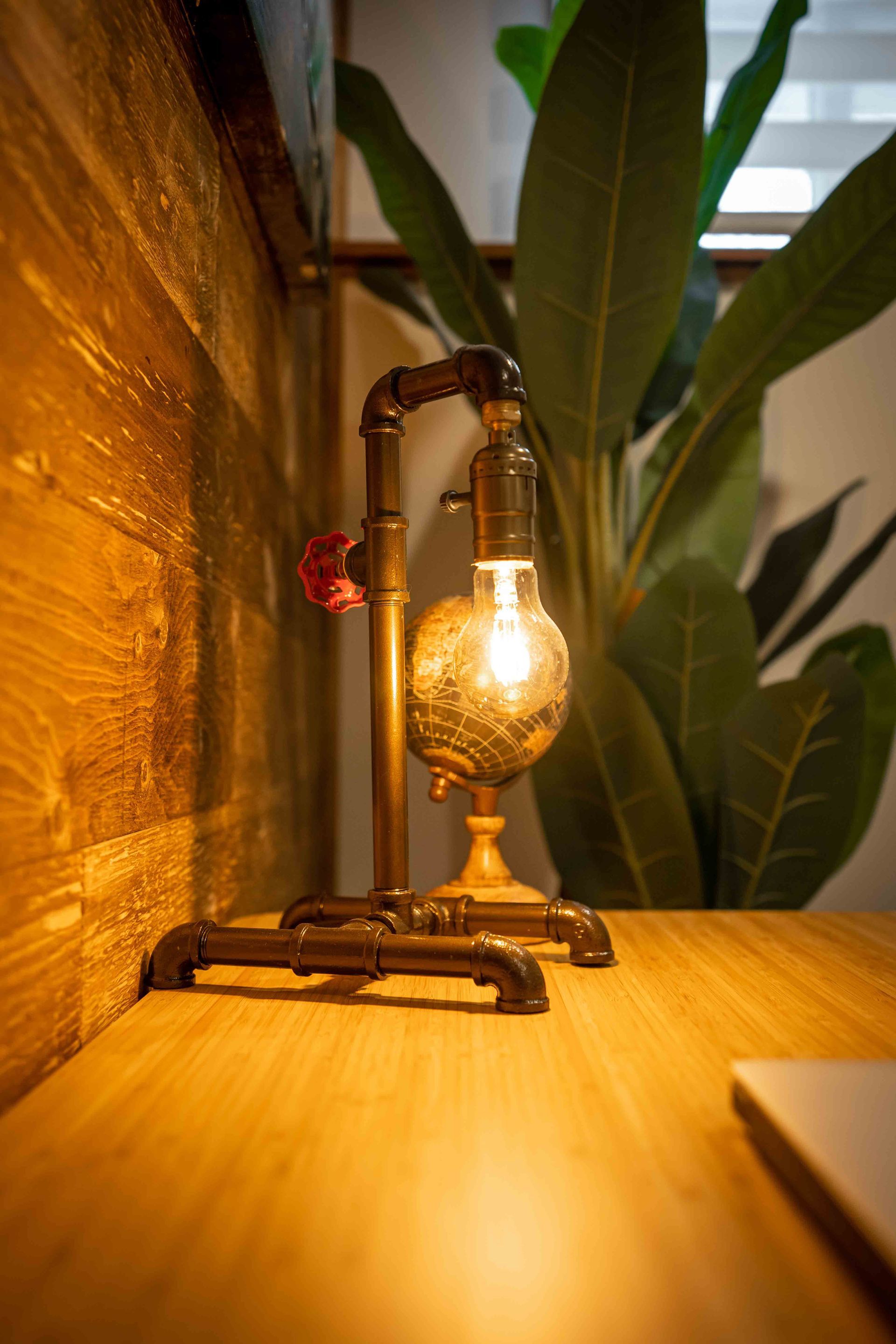 an office desk with wooden walls, a small desk top globe illuminated by a desk top vintage lamp and a large green floor plant