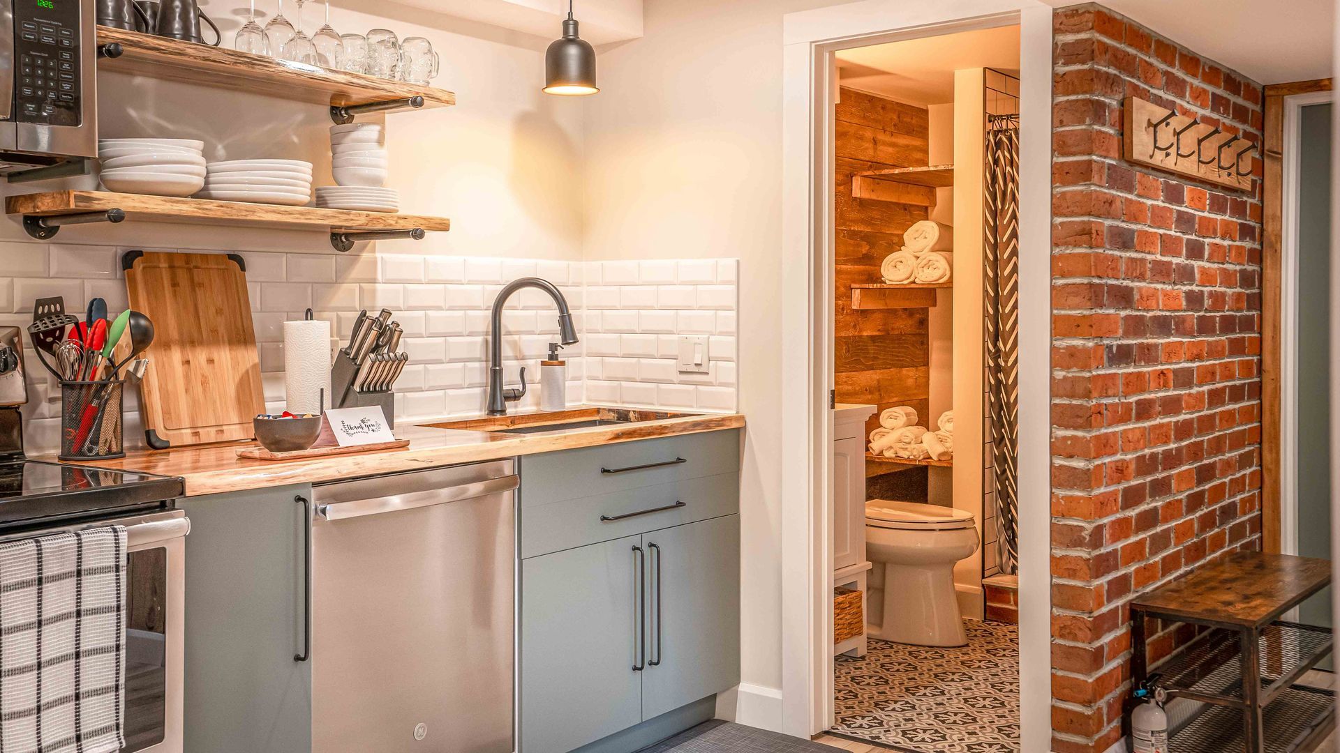 kitchen sink with dishwasher and open door to bathroom. a red brick accent wall with hooks for jackets
