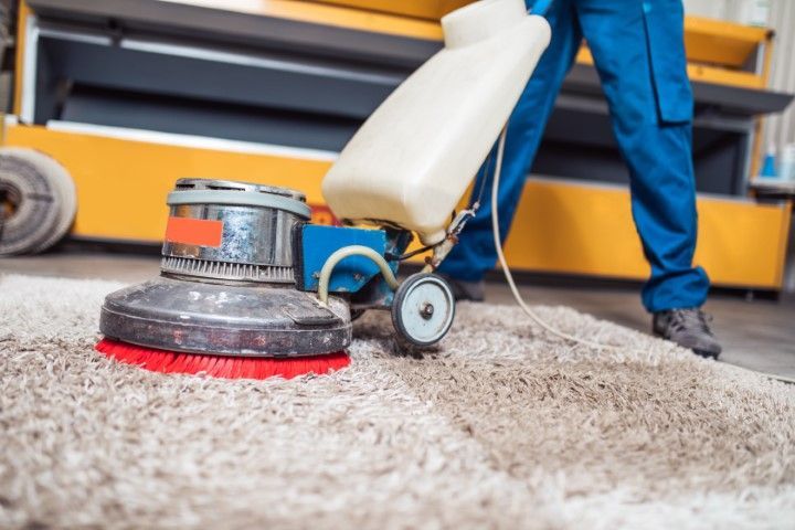 Carpet cleaner buffing a beige carpet beside a yellow truck, with a worker’s legs visible