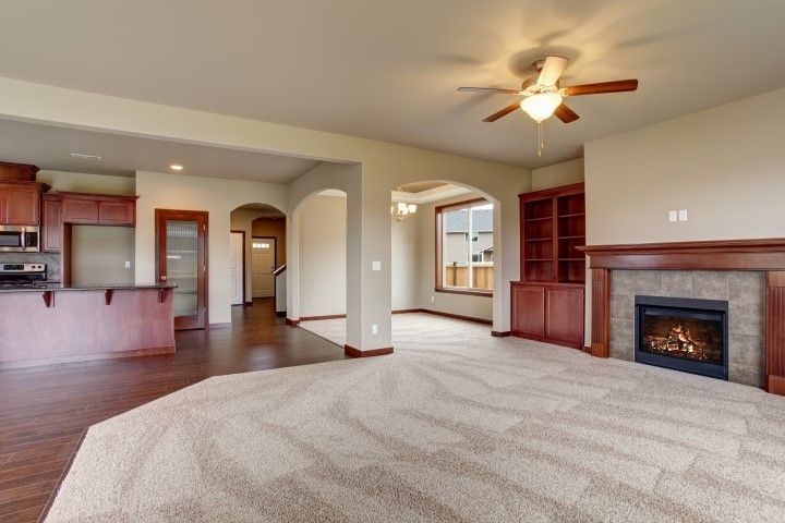 Empty living room with beige carpet, hardwood floors, fireplace, ceiling fan, and open kitchen view