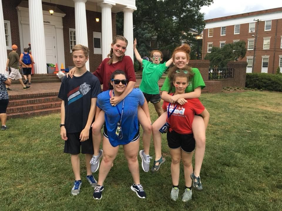 Group of kids and young adults posing outdoors on a grassy area. Some are giving piggyback rides and smiling near a white-columned building.