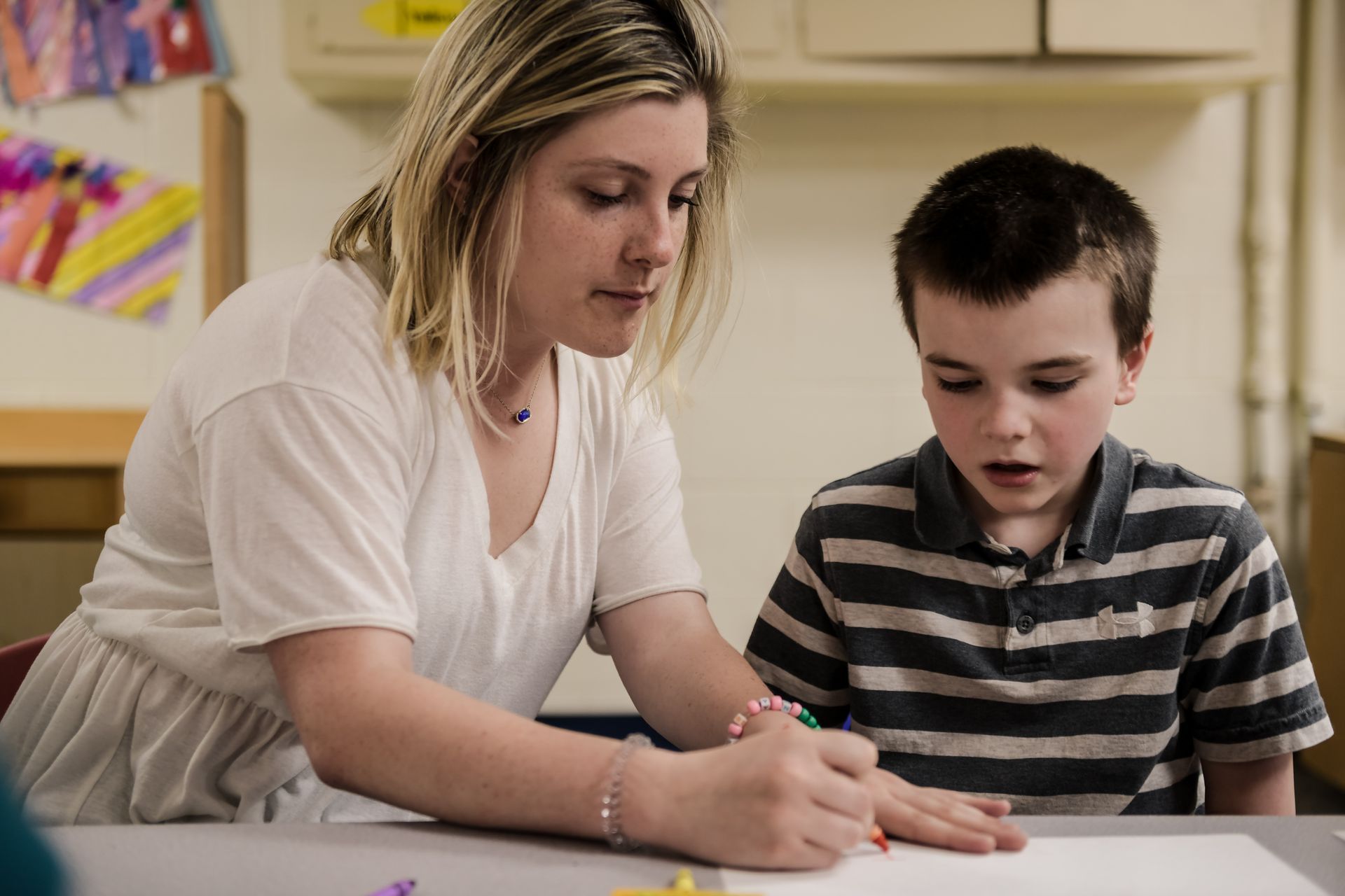 A woman and boy at a table, drawing together. The woman wears a white shirt, the boy a striped one. They appear focused in a room with art on the wall.