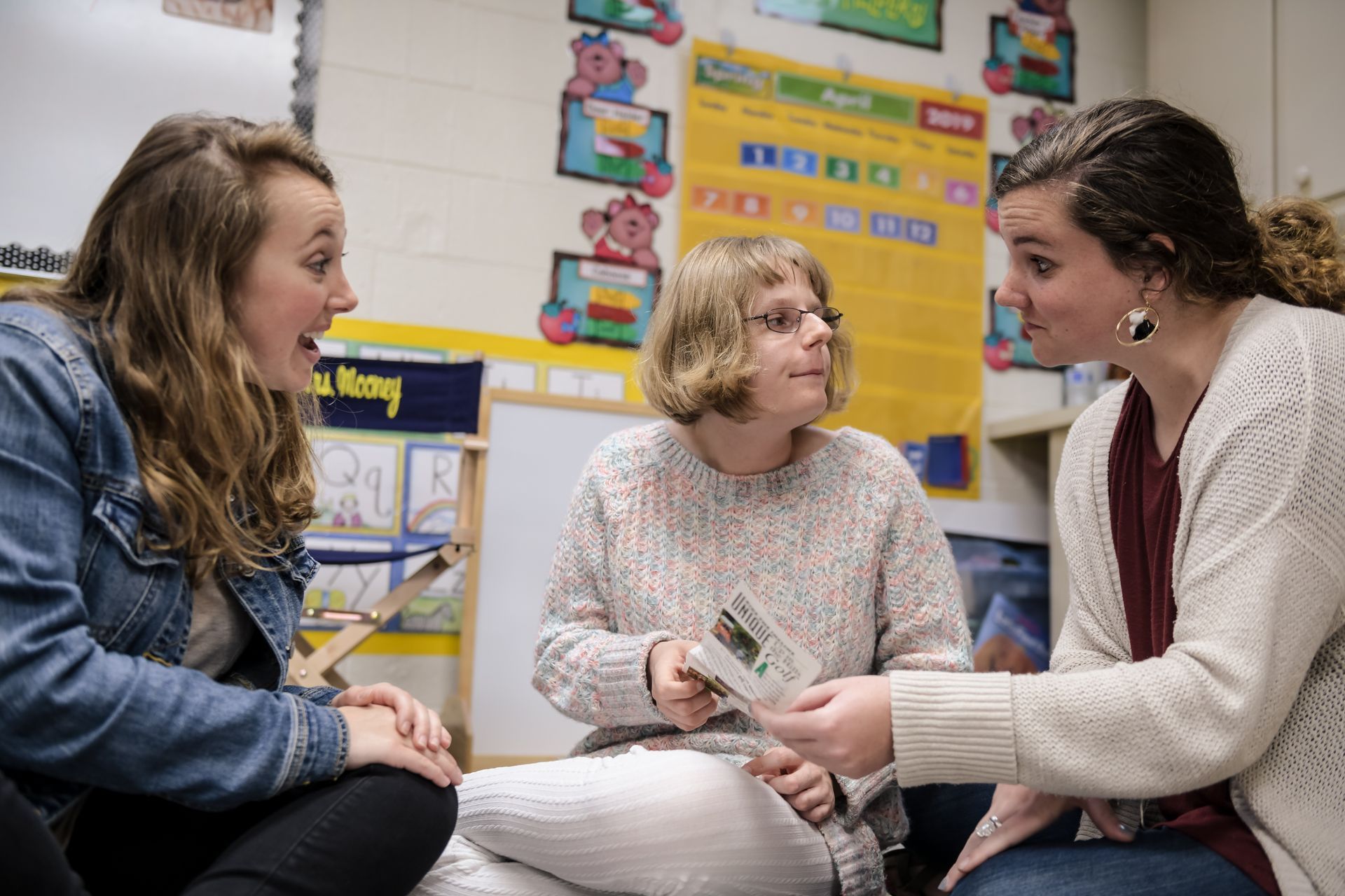 Three women in a classroom, two are looking at a book held by the woman in the middle. They appear to be having a discussion.