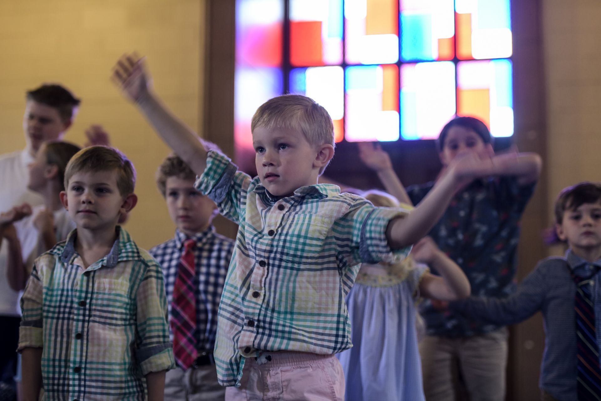 A group of young children, some wearing dress shirts and ties, with arms raised in front of a stained-glass window.